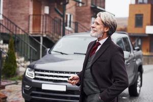 alla moda anziano uomo con grigio capelli e barba è all'aperto su il strada vicino il suo auto foto