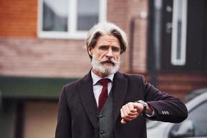alla moda anziano uomo con grigio capelli e barba è all'aperto su il strada vicino il suo auto assegni tempo su il suo orologio foto