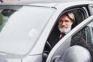 alla moda anziano uomo con grigio capelli e barba è all'aperto su il strada seduta nel il suo auto foto