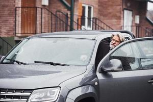 alla moda anziano uomo con grigio capelli e barba è all'aperto su il strada seduta nel il suo auto foto