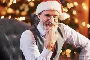 ritratto di elegante anziano con grigio capelli e barba nel giacca e bianca camicia è nel il decorato Natale camera foto