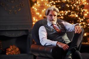 ritratto di elegante anziano con grigio capelli e barba è nel decorato Natale camera seduta su il sedia foto