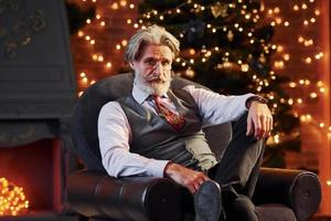 ritratto di elegante anziano con grigio capelli e barba è nel decorato Natale camera seduta su il sedia foto