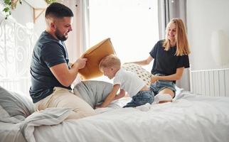 giovane sposato coppia con loro giovane figlio giocando cuscino combattimento nel Camera da letto a giorno foto