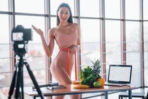 giovane fitness vlogger fare video in casa di in piedi vicino tavolo con salutare cibo e il computer portatile foto