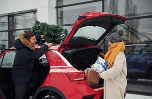 giovane coppia con regalo scatole è vicino auto con albero su il superiore. insieme all'aperto a inverno tempo foto