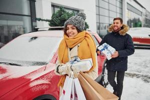 giovane coppia con regalo scatole è vicino auto con albero su il superiore. insieme all'aperto a inverno tempo foto