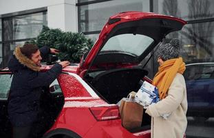 giovane coppia con regalo scatole è vicino auto con albero su il superiore. insieme all'aperto a inverno tempo foto