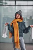 sorridente e sensazione contento. bellissimo allegro ragazza nel giallo sciarpa e nel caldo Abiti in piedi in casa con shopping borse nel mani foto