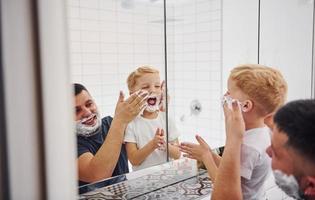 padre con il suo figlio è nel il bagno avere divertimento di utilizzando rasatura gel e guardare nel il specchio foto