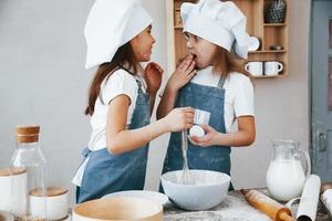 Due poco ragazze nel blu capocuoco uniforme parlando segreti per ogni altro quando preparazione cibo su il cucina foto