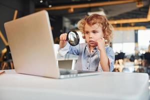 inteligente bambino nel casuale Abiti con il computer portatile su tavolo avere divertimento con ingrandimento bicchiere foto