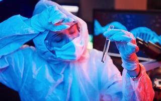 infermiera nel maschera e bianca uniforme, Tenere tubo con liquido e seduta nel neon illuminato laboratorio con computer e medico attrezzatura ricerca per coronavirus vaccino foto