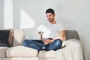 uomo nel bianca camicia e jeans seduta su letto con il computer portatile in casa foto