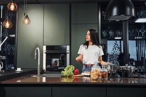 preparazione cibo di utilizzando verdure e salutare prodotti. giovane bellissimo brunetta nel casuale Abiti in casa nel cucina a giorno foto