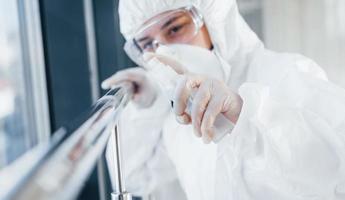 femmina medico scienziato nel laboratorio cappotto, difensiva occhiali e maschera in piedi in casa con antibatterico spray foto