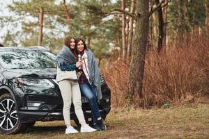 madre e figlia in piedi insieme vicino moderno nero auto all'aperto nel il foresta foto