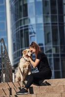 giovane positivo donna seduta su le scale con sua cane quando avere un' camminare all'aperto vicino attività commerciale edificio foto