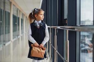 giovane poco scuola ragazza nel uniforme in piedi nel corridoio con pacchetto di cena nel mani foto