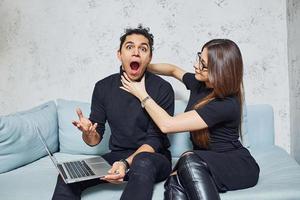 uomo con Riccio capelli e donna quello nel nero Abiti seduta con il computer portatile in casa e avere divertimento foto