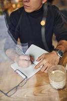 giovane maschio scrittore con nero Riccio capelli seduta in casa nel bar con penna e bloc notes. Visualizza attraverso il bicchiere foto