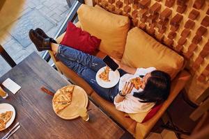 giovane brunetta seduta in casa con Pizza e smartphone nel mano foto