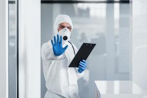 Spettacoli fermare gesto di mano. maschio medico scienziato nel laboratorio cappotto, difensiva occhiali e maschera Tenere bloc notes nel mani foto