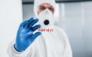 maschio medico scienziato nel laboratorio cappotto, difensiva occhiali e maschera detiene bicchiere con covid-19 parola su esso foto