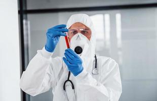 maschio medico scienziato nel laboratorio cappotto, difensiva occhiali e maschera detiene test tubo con sangue foto