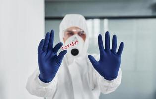 maschio medico scienziato nel laboratorio cappotto, difensiva occhiali e maschera detiene bicchiere con covid-19 parola su esso foto