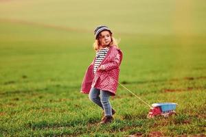 carino poco ragazza passeggiate con giocattolo auto su il bellissimo campo a soleggiato giorno foto