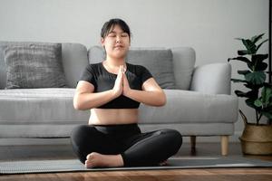 donna paffuta asiatica seduta sul pavimento in soggiorno pratica lezione di yoga. femmina che ha meditato classe di formazione. foto