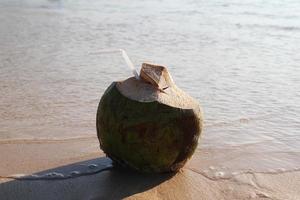 un' Noce di cocco con cannuccia su il sabbioso spiaggia vicino per il mare nel il soleggiato tempo atmosferico. foto