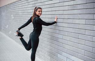giovane allegro ragazza nel nero abbigliamento sportivo all'aperto fare allungamento vicino grigio parete foto