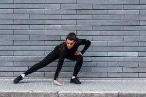 giovane allegro ragazza nel nero abbigliamento sportivo all'aperto fare allungamento vicino grigio parete foto