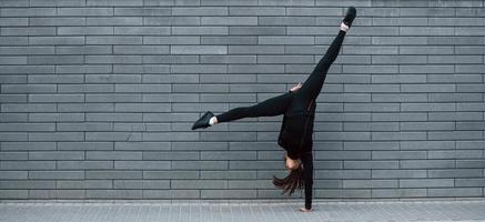 giovane allegro ragazza nel nero abbigliamento sportivo fare difficile handstand esercizi all'aperto vicino grigio parete foto