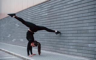 giovane allegro ragazza nel nero abbigliamento sportivo fare difficile handstand esercizi all'aperto vicino grigio parete foto