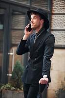 parlando di Telefono. elegante giovane uomo nel nero cappello e Abiti con ombrello nel mani all'aperto nel il città foto