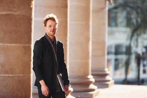 con il computer portatile nel mani. elegante giovane uomo nel bicchieri adn nel formale di classe Abiti all'aperto nel il città foto