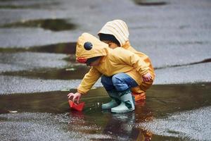 Due bambini nel giallo impermeabile mantelli e stivali giocando con fatto a mano carta barca all'aperto dopo il pioggia insieme foto