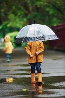 Due bambini con ombrello nel giallo impermeabile mantelli e stivali giocando all'aperto dopo il pioggia insieme foto