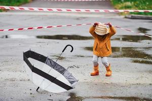 ragazzo nel giallo impermeabile mantello, stivali e con ombrello giocando all'aperto vicino protettivo nastro dopo il pioggia foto