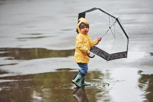 ragazzo nel giallo impermeabile mantello, stivali e con ombrello giocando all'aperto dopo il pioggia foto