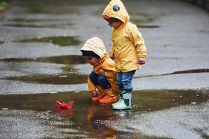 Due bambini nel giallo impermeabile mantelli e stivali giocando con fatto a mano carta barca all'aperto dopo il pioggia insieme foto