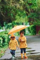 Due bambini con ombrello nel giallo impermeabile mantelli e stivali giocando all'aperto dopo il pioggia insieme foto