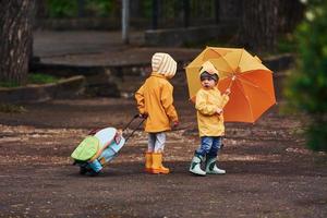 Due bambini con ombrello, valigia e giallo impermeabile mantelli e stivali a piedi all'aperto dopo il pioggia insieme foto