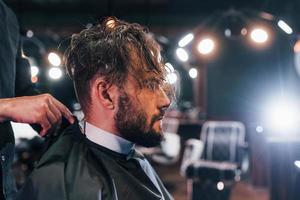 giovane barbuto uomo seduta e ottenere taglio di capelli nel barbiere negozio foto