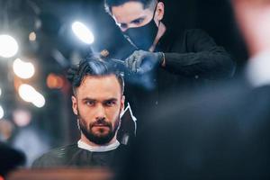 davanti Visualizza di giovane barbuto uomo quello seduta e ottenere taglio di capelli nel barbiere negozio di tipo nel nero protettivo maschera foto