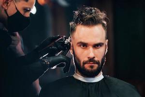 davanti Visualizza di giovane barbuto uomo quello seduta e ottenere taglio di capelli nel barbiere negozio di tipo nel nero protettivo maschera foto