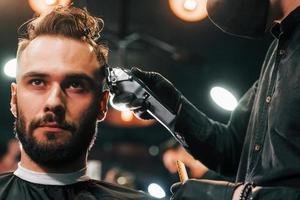 davanti Visualizza di giovane barbuto uomo quello seduta e ottenere taglio di capelli nel barbiere negozio di tipo nel nero protettivo maschera foto